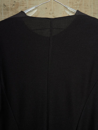 A close-up of a black knit top hanging on a clear plastic hanger against a light beige textured wall.