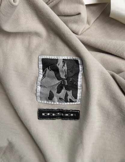 A close-up of the beige fabric from the Rick Owens DRKSHDW AW18 “SISYPHUS” Pearl Racer Tank Top shows two stitched labels: one with a black and white abstract design, the other featuring DREWSHOW in white on black.