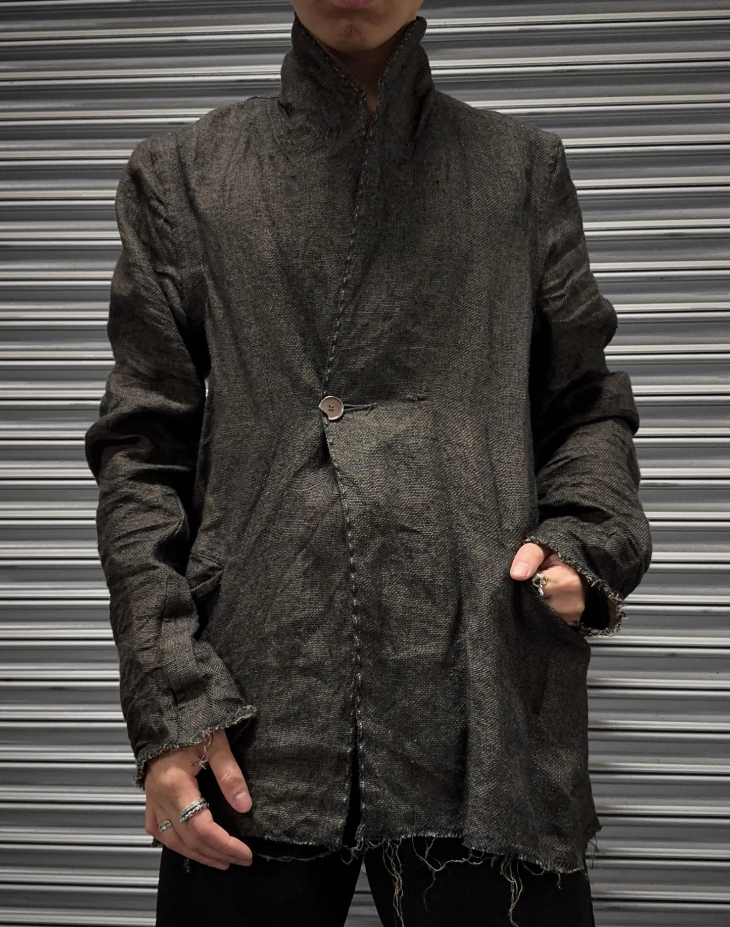 A person wears the TaichiMurakami “Coin” Taped Seam Collarless Work Blazer—a dark, textured Japanese linen jacket with frayed edges and one button—standing before a metal shutter. The photo is cropped at the neck, hand in pocket.