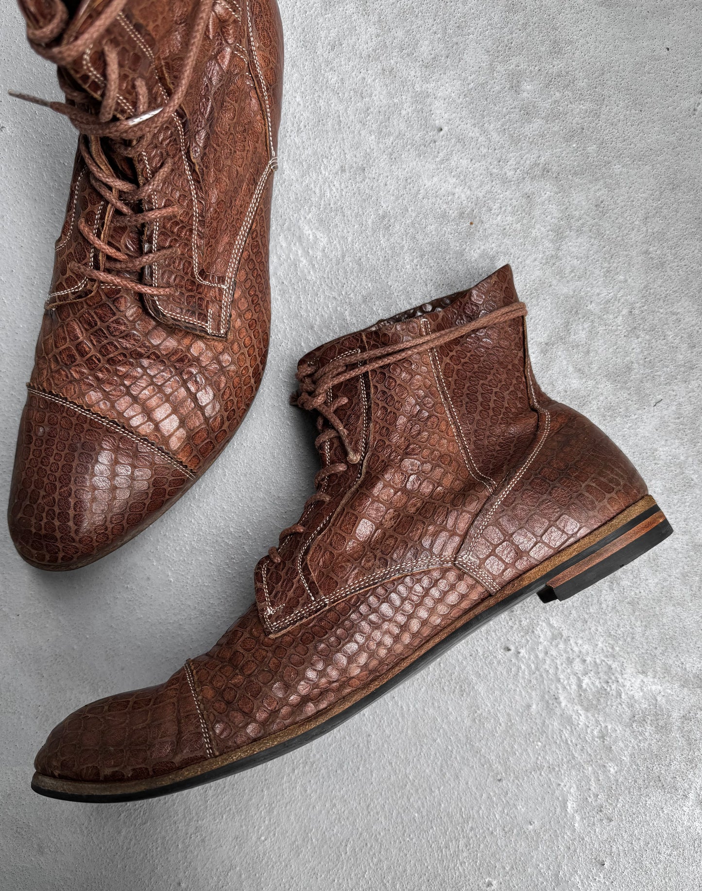 Mihara Yasuhiro Early 00s Python Embossed Cow Leather Pointy Toe Boots, featuring a brown, lace-up design with python texture, are inspired by avant-garde fashion and styled on a light gray concrete surface.