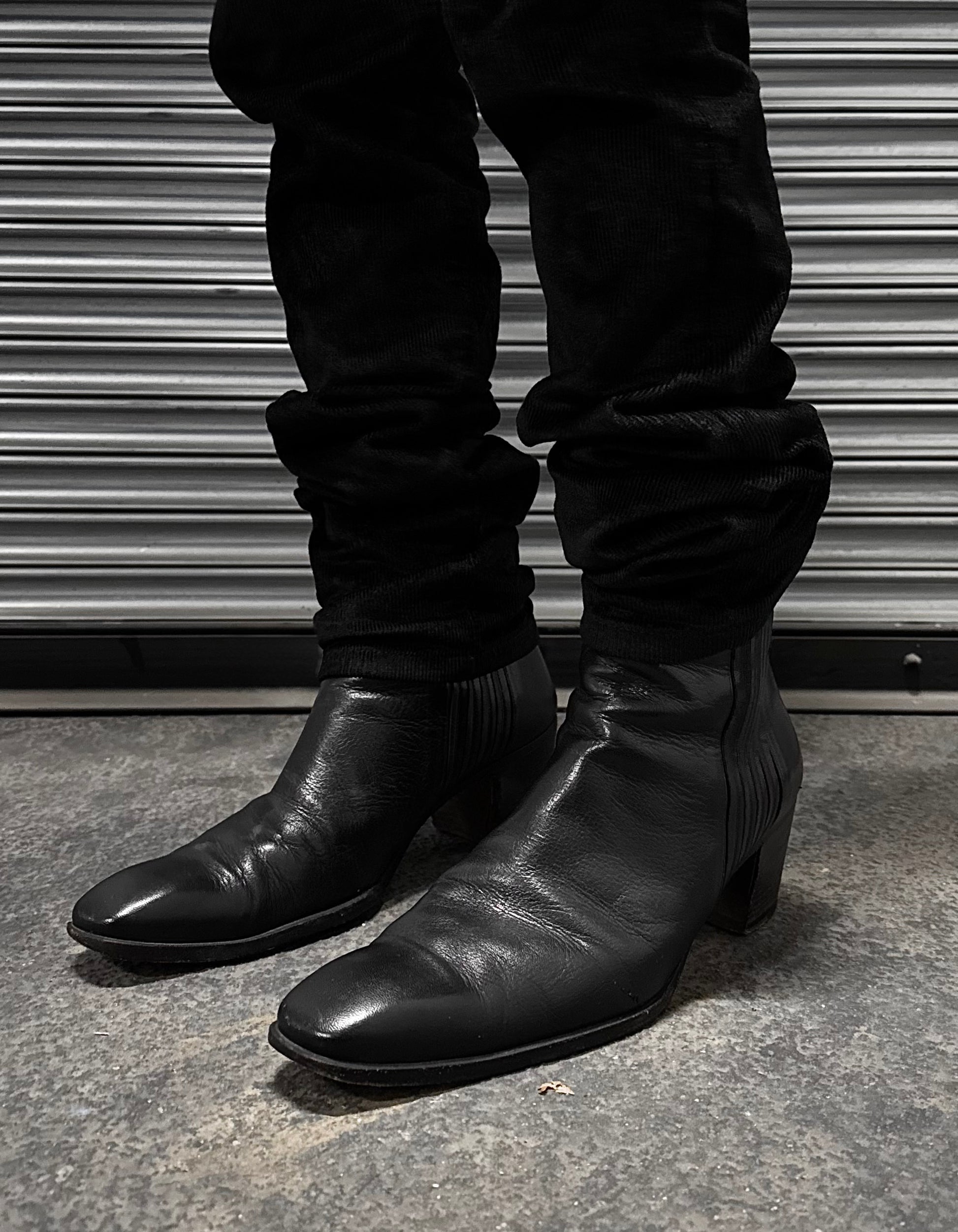 A person in fitted black pants and Saint Laurent Paris Rive Gauche SS04 by Tom Ford 40mm Heel ‘Johnny’ Leather Boots stands on a concrete floor before a metallic ribbed shutter.
