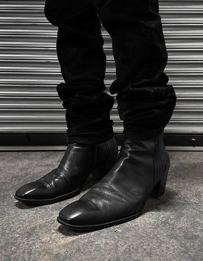 A person in fitted black pants and Saint Laurent Paris Rive Gauche SS04 by Tom Ford 40mm Heel ‘Johnny’ Leather Boots stands on a concrete floor before a metallic ribbed shutter.