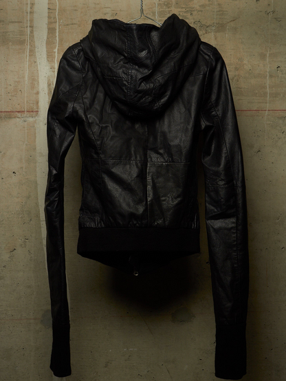 A black faux-leather hooded jacket with long sleeves, shown from the back, hanging on a metal hanger against a textured concrete wall.