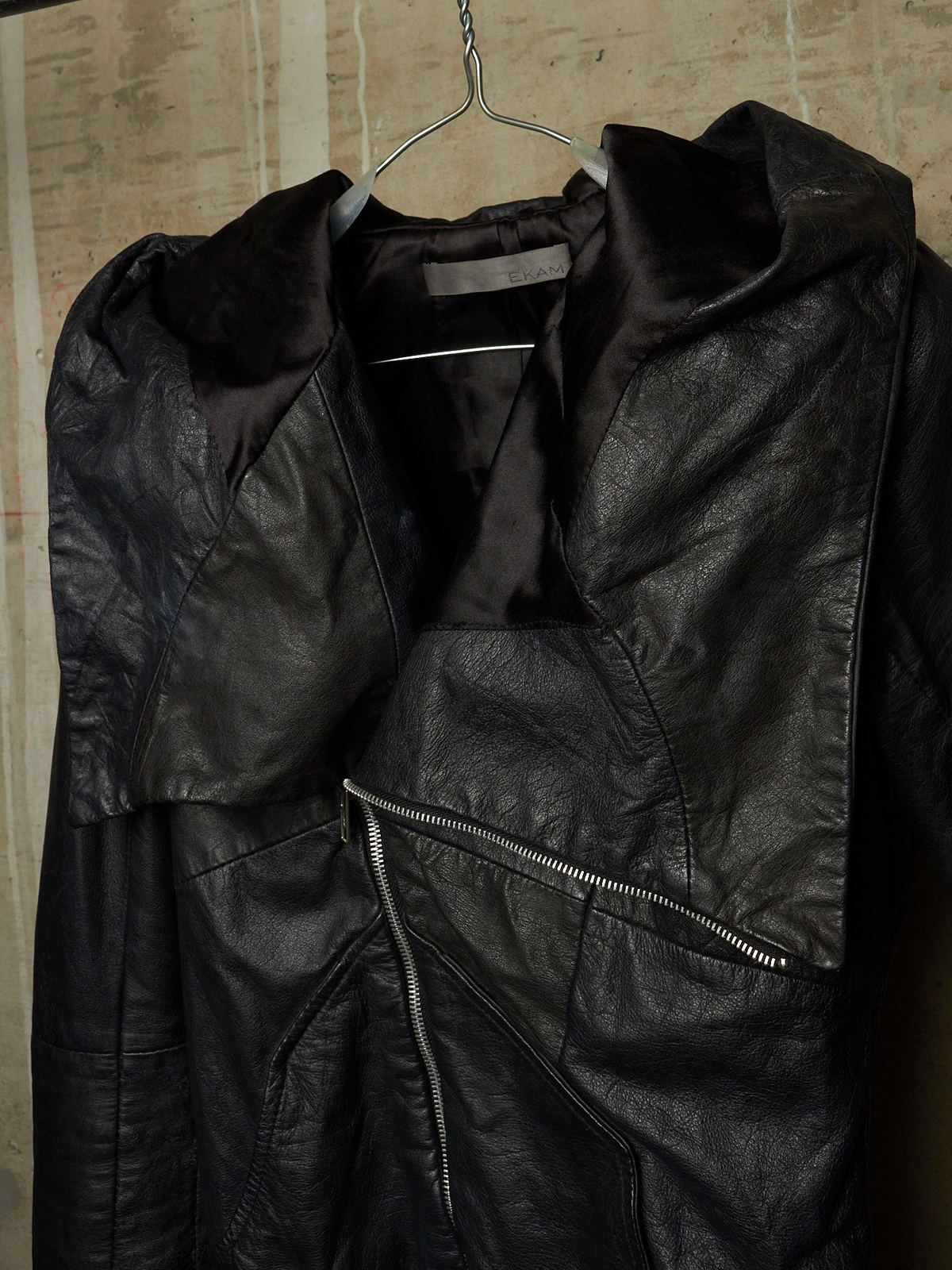 A black leather jacket with a high collar and asymmetrical zipper, hanging on a metal hanger against a textured beige wall.