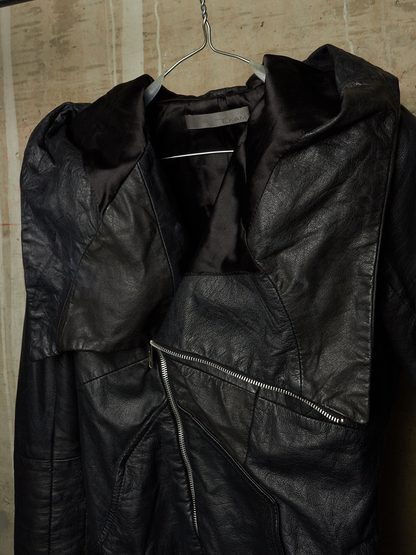 A black leather jacket with a high collar and asymmetrical zipper, hanging on a metal hanger against a textured beige wall.