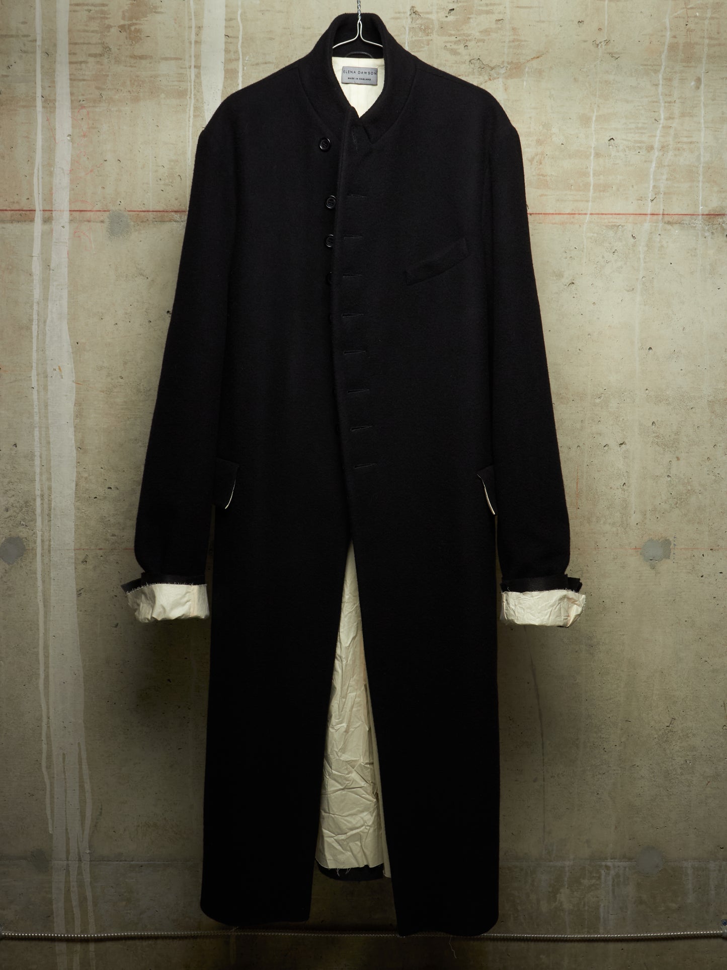 A long black coat with a high collar and front buttons, hanging against a textured concrete wall. The coat has white cuffs and a white lining visible at the opening.