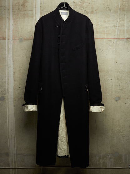 A long black coat with a high collar and front buttons, hanging against a textured concrete wall. The coat has white cuffs and a white lining visible at the opening.