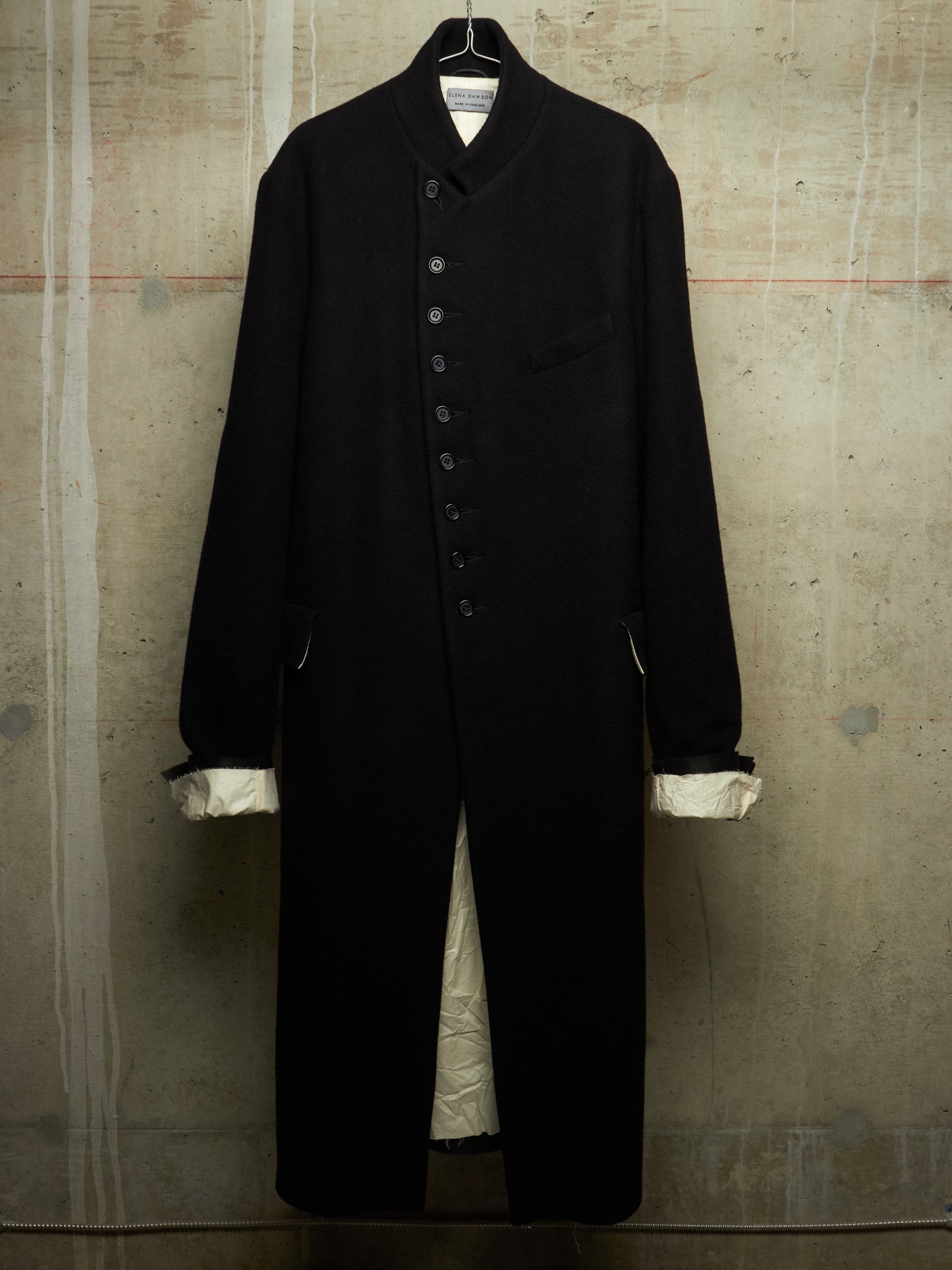 A long black wool-blend coat with a high stand collar, front button closure, and chest pocket, hanging against a textured concrete wall. The cuffs are turned up to show a contrasting cream lining.