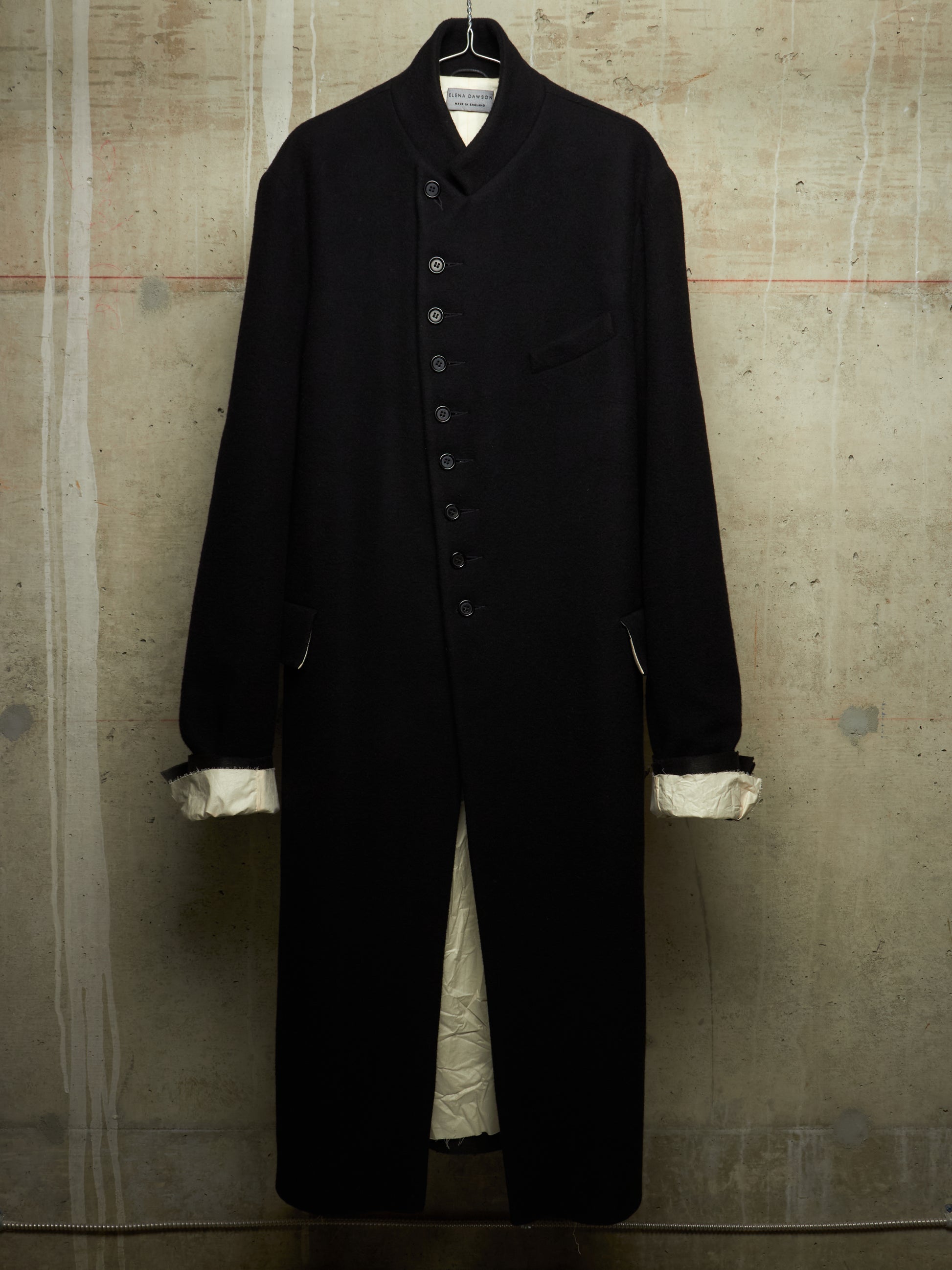 A long black wool-blend coat with a high stand collar, front button closure, and chest pocket, hanging against a textured concrete wall. The cuffs are turned up to show a contrasting cream lining.