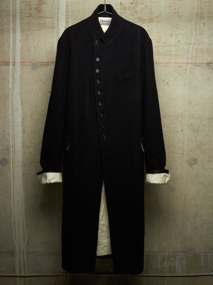 A long black wool-blend coat with a high stand collar, front button closure, and chest pocket, hanging against a textured concrete wall. The cuffs are turned up to show a contrasting cream lining.