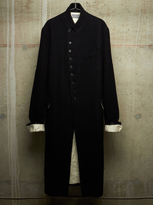 A long black wool-blend coat with a high stand collar, front button closure, and chest pocket, hanging against a textured concrete wall. The cuffs are turned up to show a contrasting cream lining.