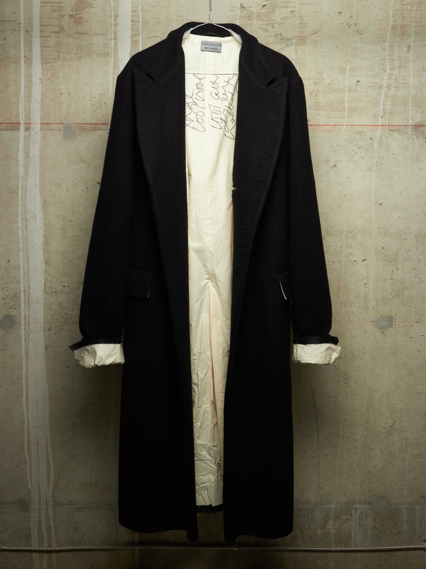 A long black coat hanging over a cream-colored dress or tunic with embroidered detailing, set against a textured concrete wall.