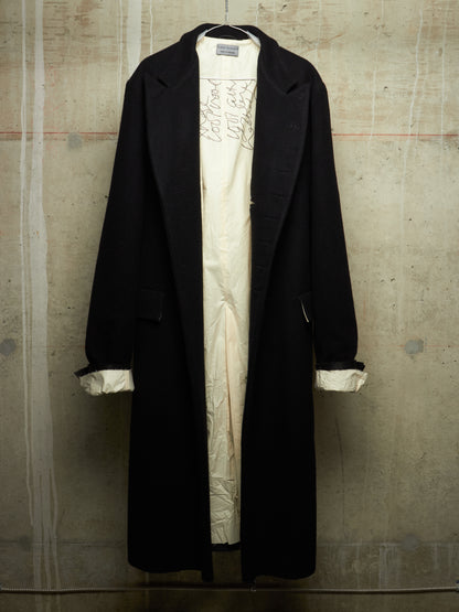 A long black coat hanging over a cream-colored dress or tunic with embroidered detailing, set against a textured concrete wall.