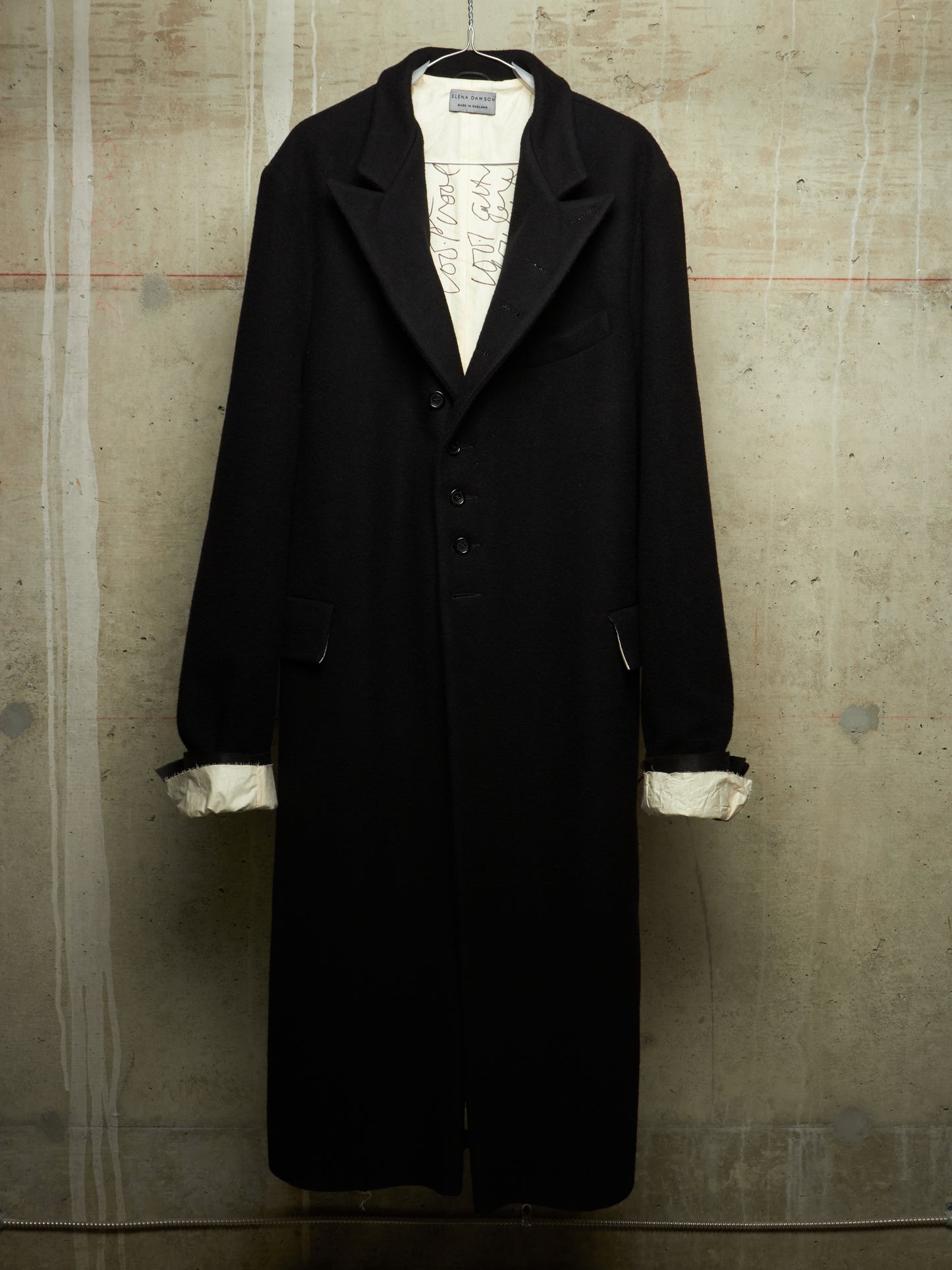 A long black coat with wide lapels and a three-button front, displayed on a hanger against a textured concrete wall. The coat is styled over a white shirt with rolled cuffs showing.