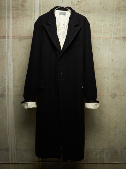 A long black coat with wide lapels and a three-button front, displayed on a hanger against a textured concrete wall. The coat is styled over a white shirt with rolled cuffs showing.