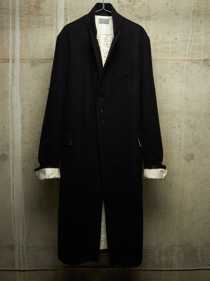 A long black coat hanging against a textured concrete wall, with white shirt cuffs visible at the sleeves and a white patterned inner layer peeking out at the front opening.