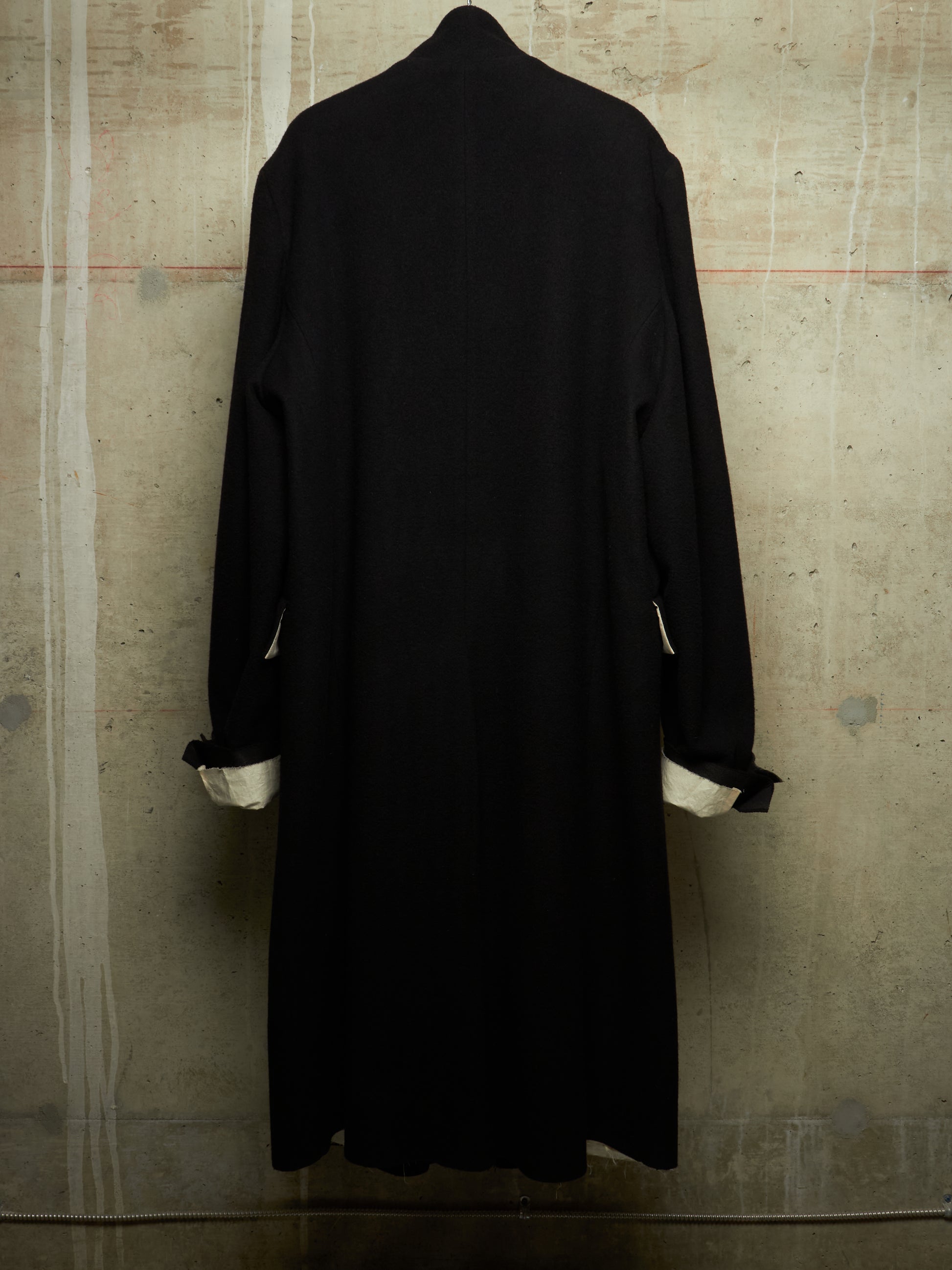 A long black coat or duster with rolled cuffs showing a white lining, displayed from the back against a textured concrete wall.