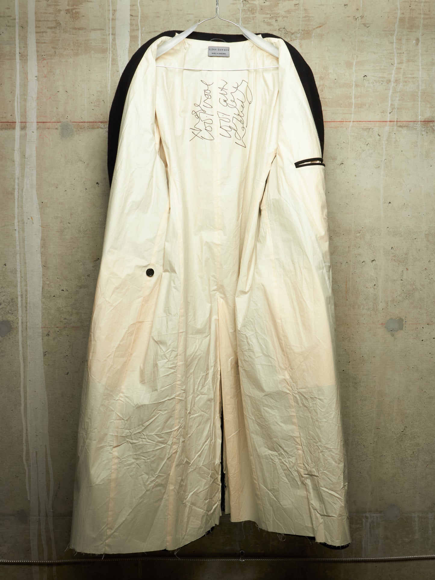 A long cream-colored coat with black shoulder panels and black trim, hanging against a textured concrete wall. The coat features hand-drawn style markings on the chest and a small black button on the side.