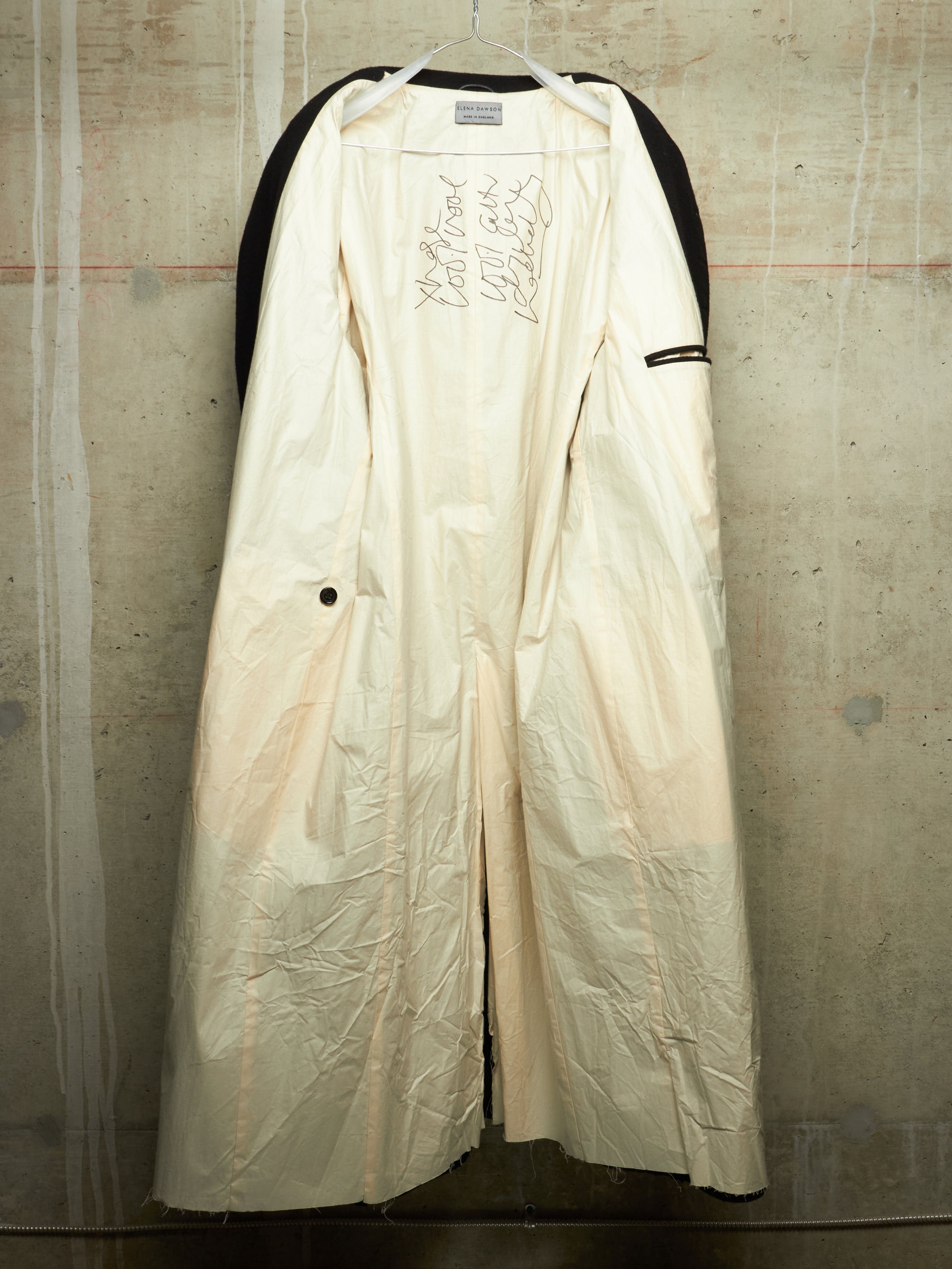 A long cream-colored coat with black shoulder panels and black trim, hanging against a textured concrete wall. The coat features hand-drawn style markings on the chest and a small black button on the side.