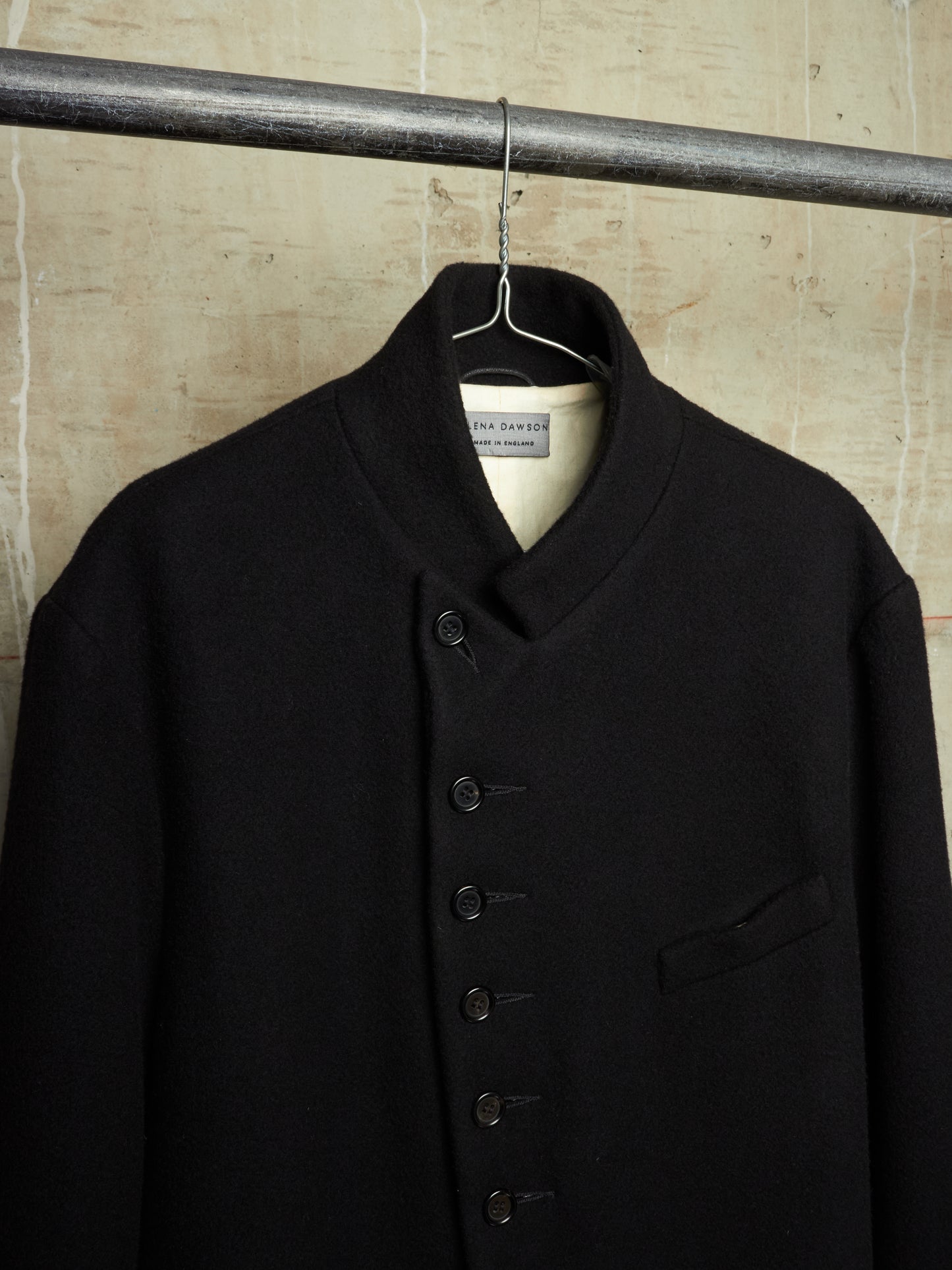 A black wool-blend jacket with a high stand collar and button closure, hanging on a metal hanger from a horizontal rod against a textured beige wall.