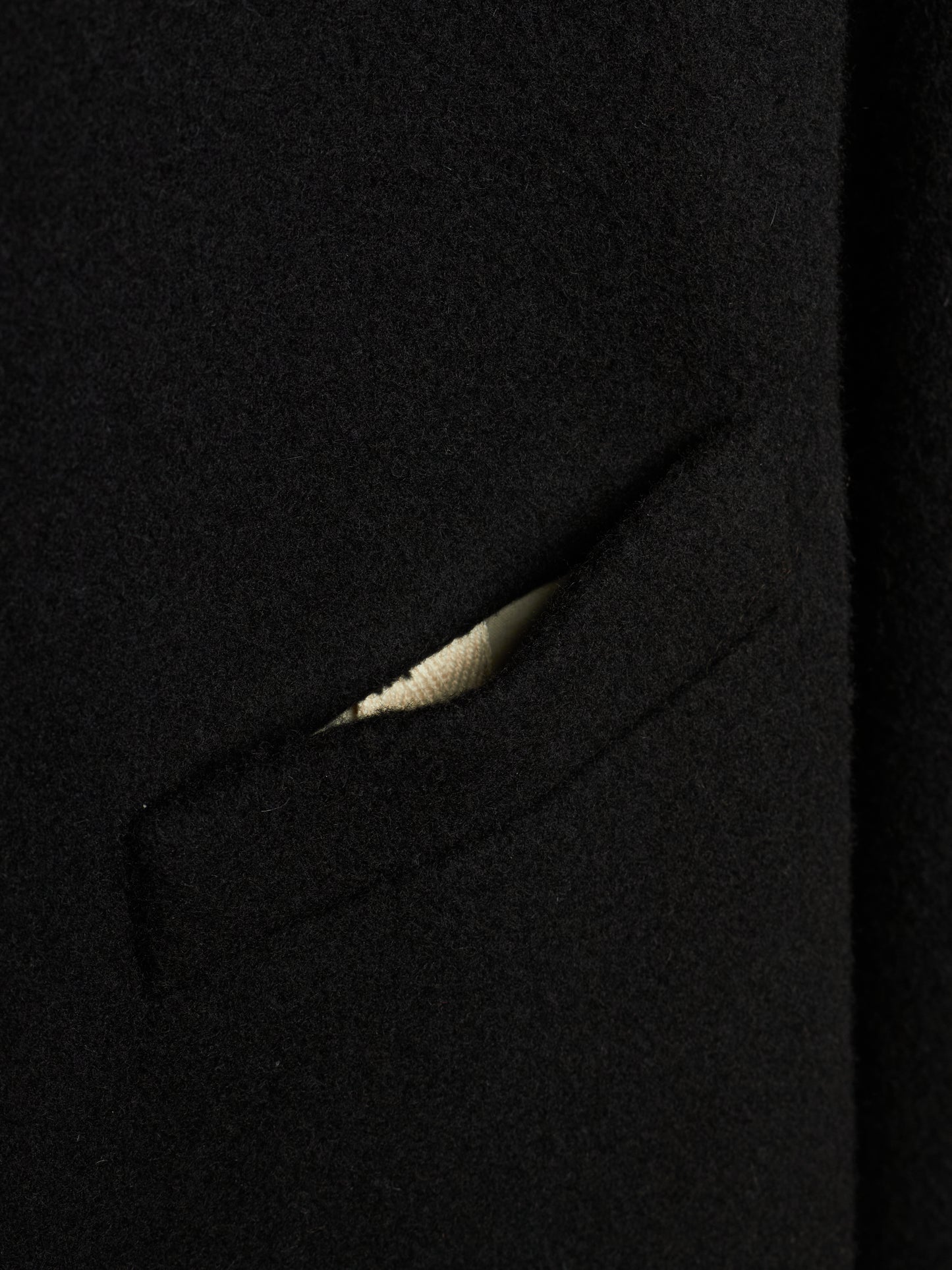 Close-up detail of a black wool or cashmere fabric garment, showing a sliver of a light beige or cream ribbed knit lining or pocket detail.