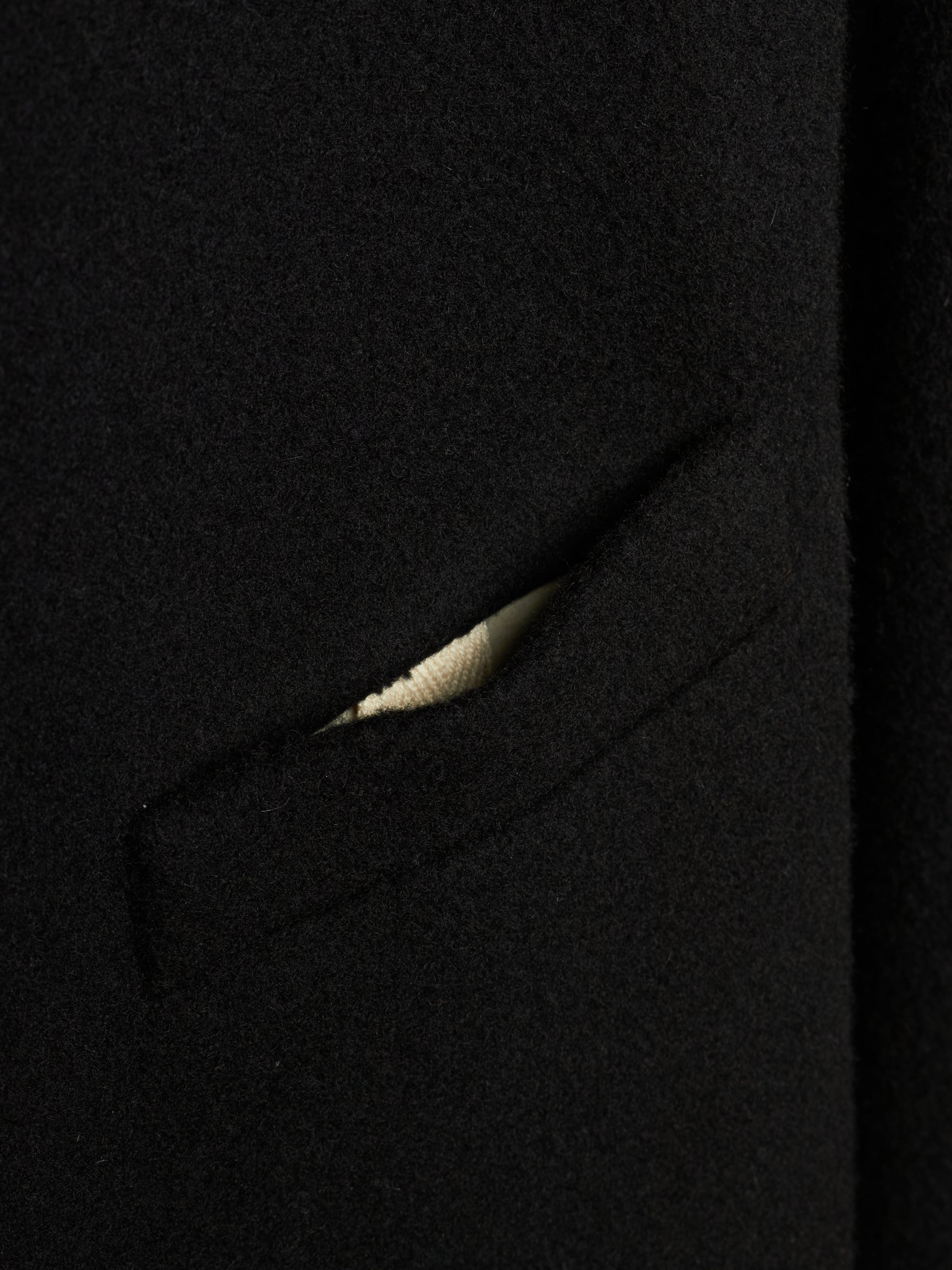 Close-up detail of a black wool or cashmere fabric garment, showing a sliver of a light beige or cream ribbed knit lining or pocket detail.