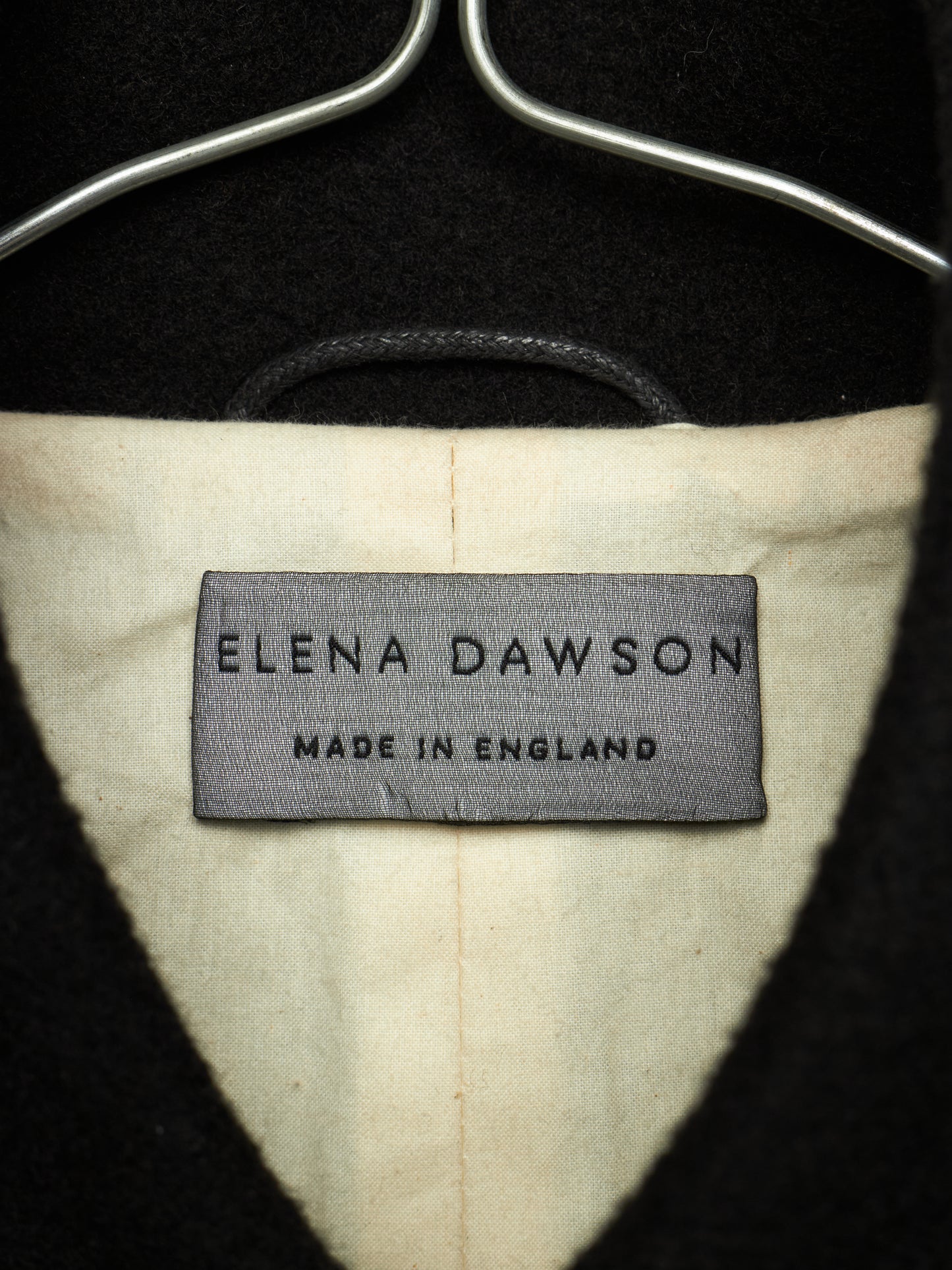 Close-up of the interior neck label of a black coat or sweater on a hanger, showing the brand 'Elena Dawson' and 'Made in England' on a cream-colored lining.