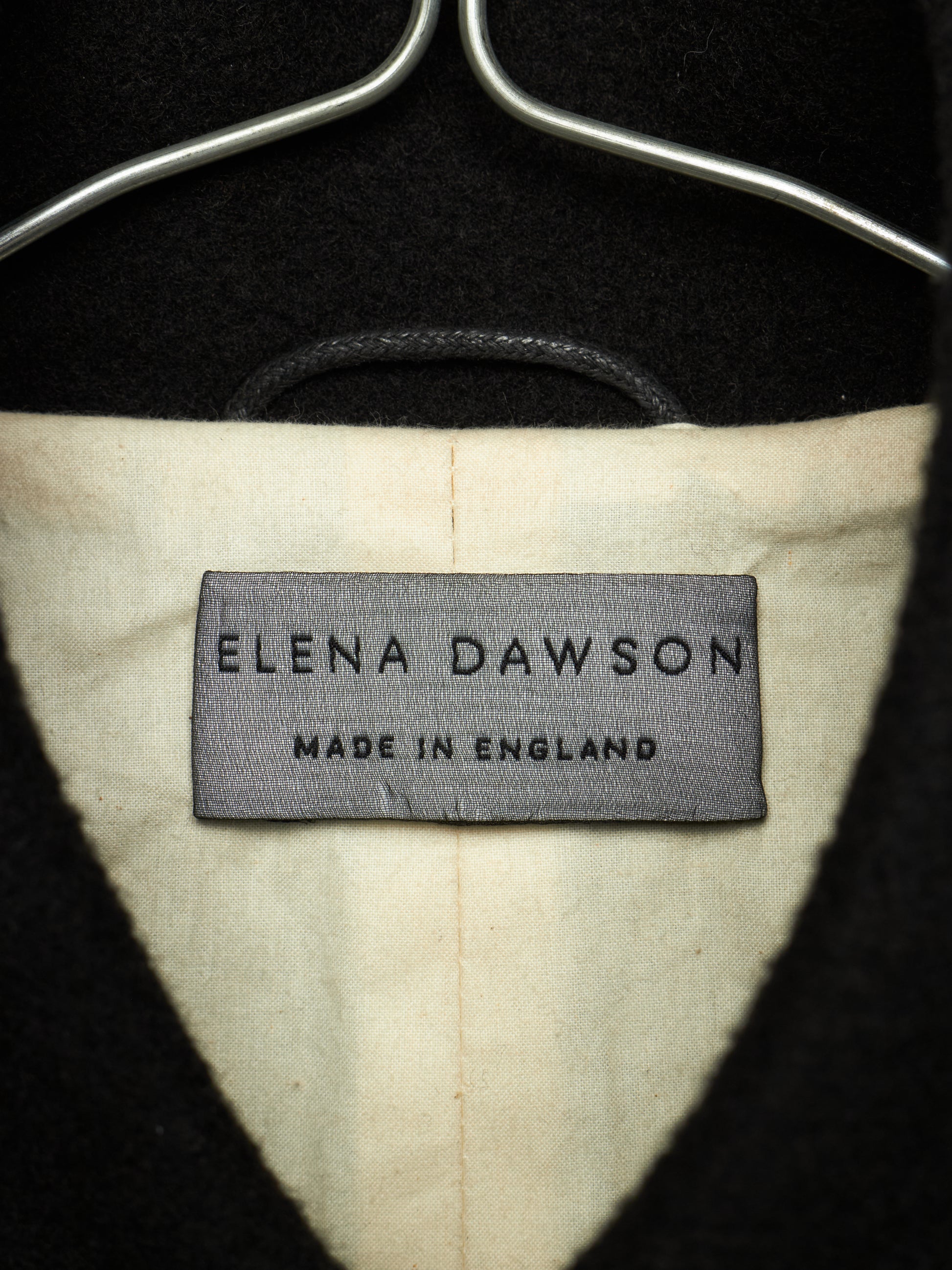 Close-up of the interior neck label of a black coat or sweater on a hanger, showing the brand 'Elena Dawson' and 'Made in England' on a cream-colored lining.