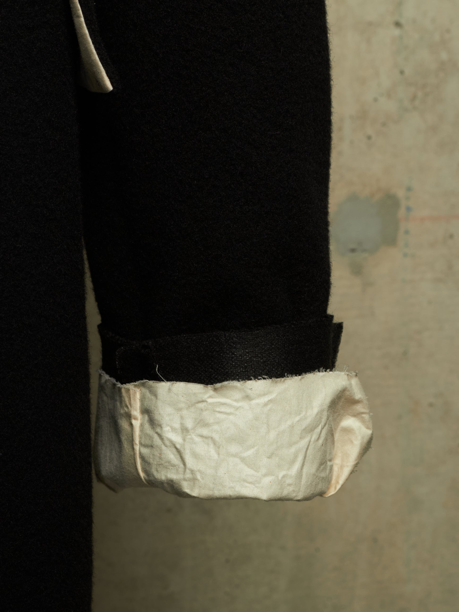 A close-up of a black coat sleeve with a rolled cuff showing a textured off-white lining, set against a neutral concrete wall.