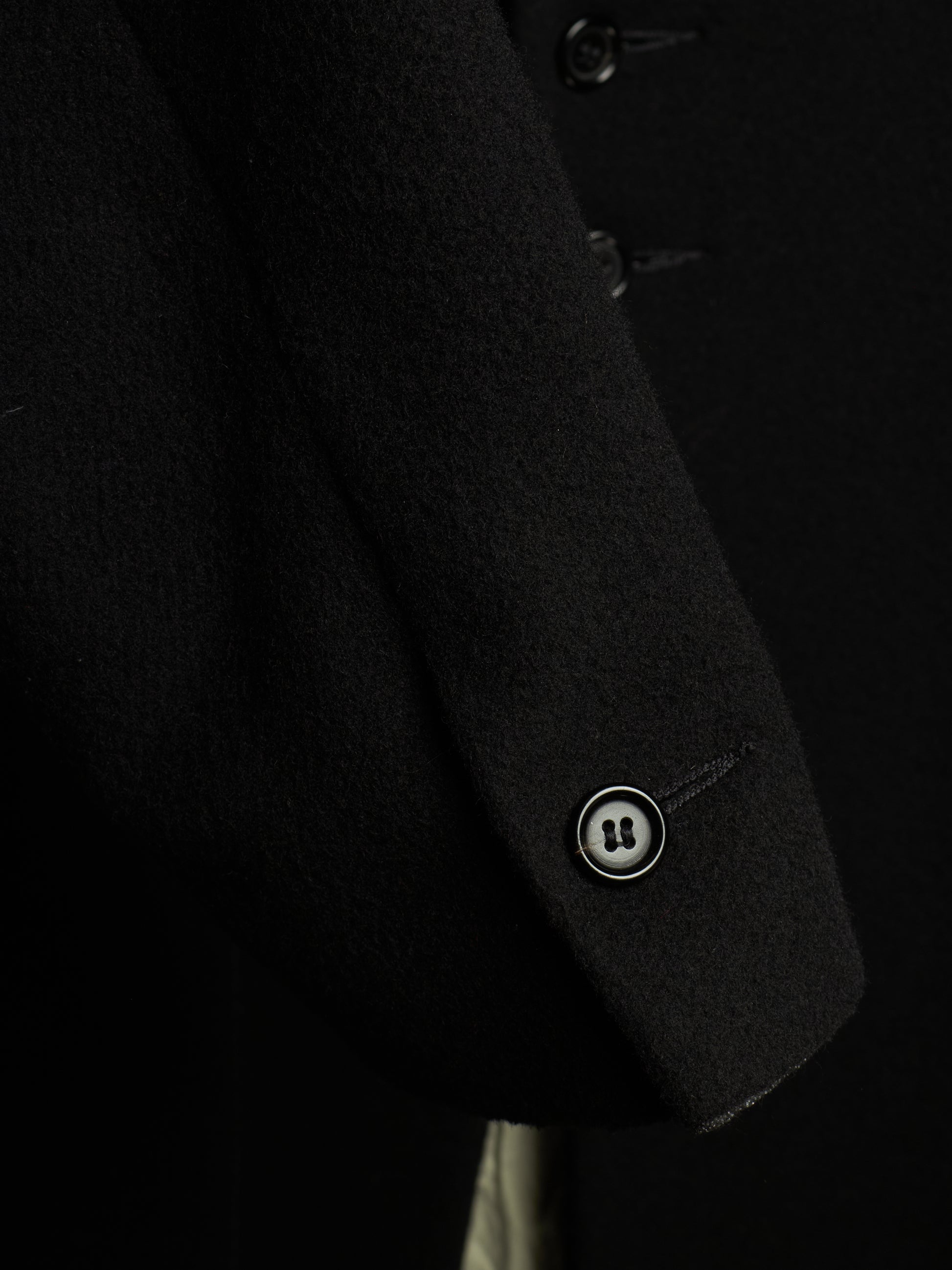 Close-up of a black wool-blend coat sleeve showing a single buttoned cuff with a silver-toned button and a small embroidered logo.