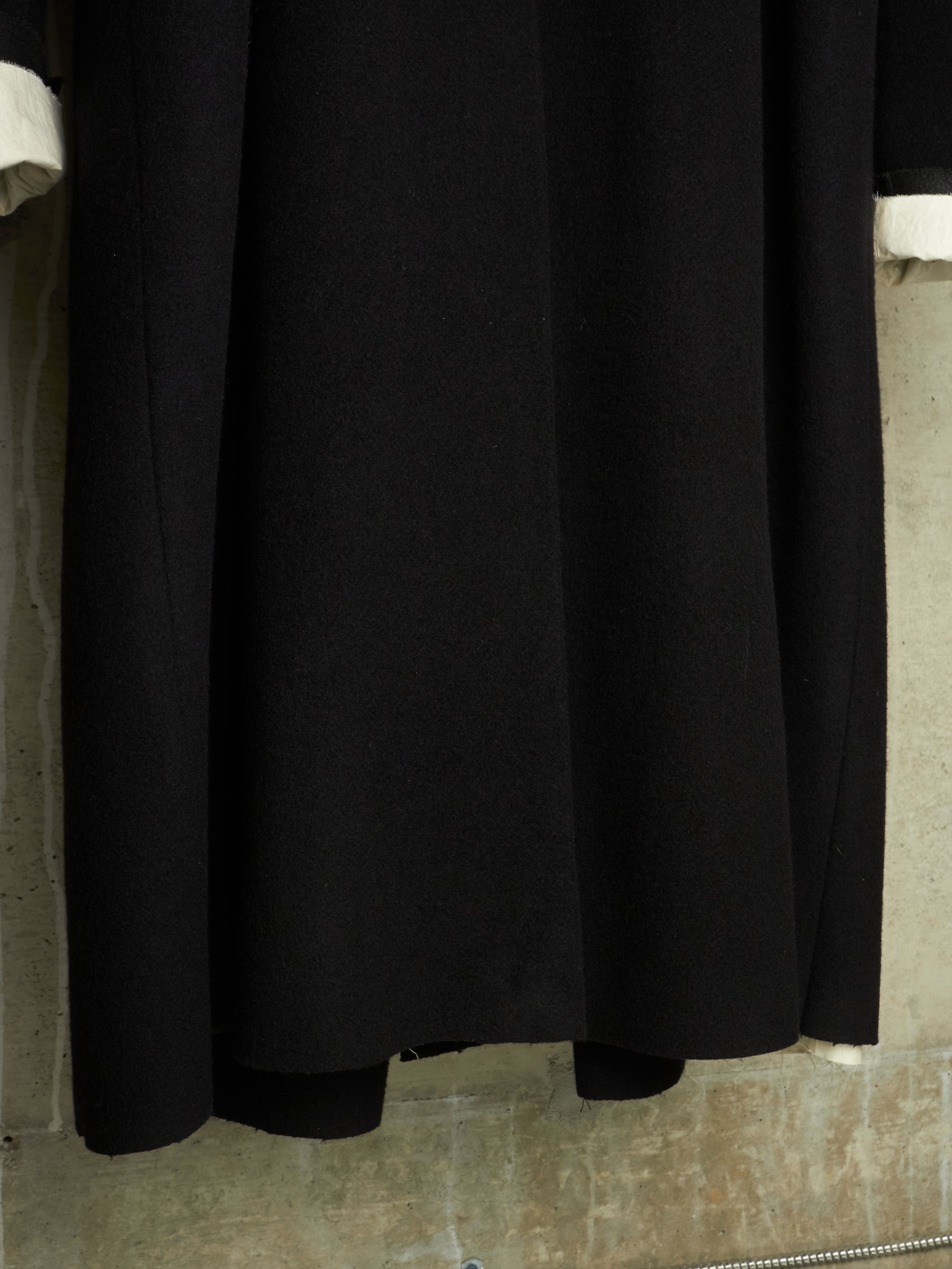 A close-up of a black long-sleeve dress or coat with white cuffs, hanging against a textured beige wall.
