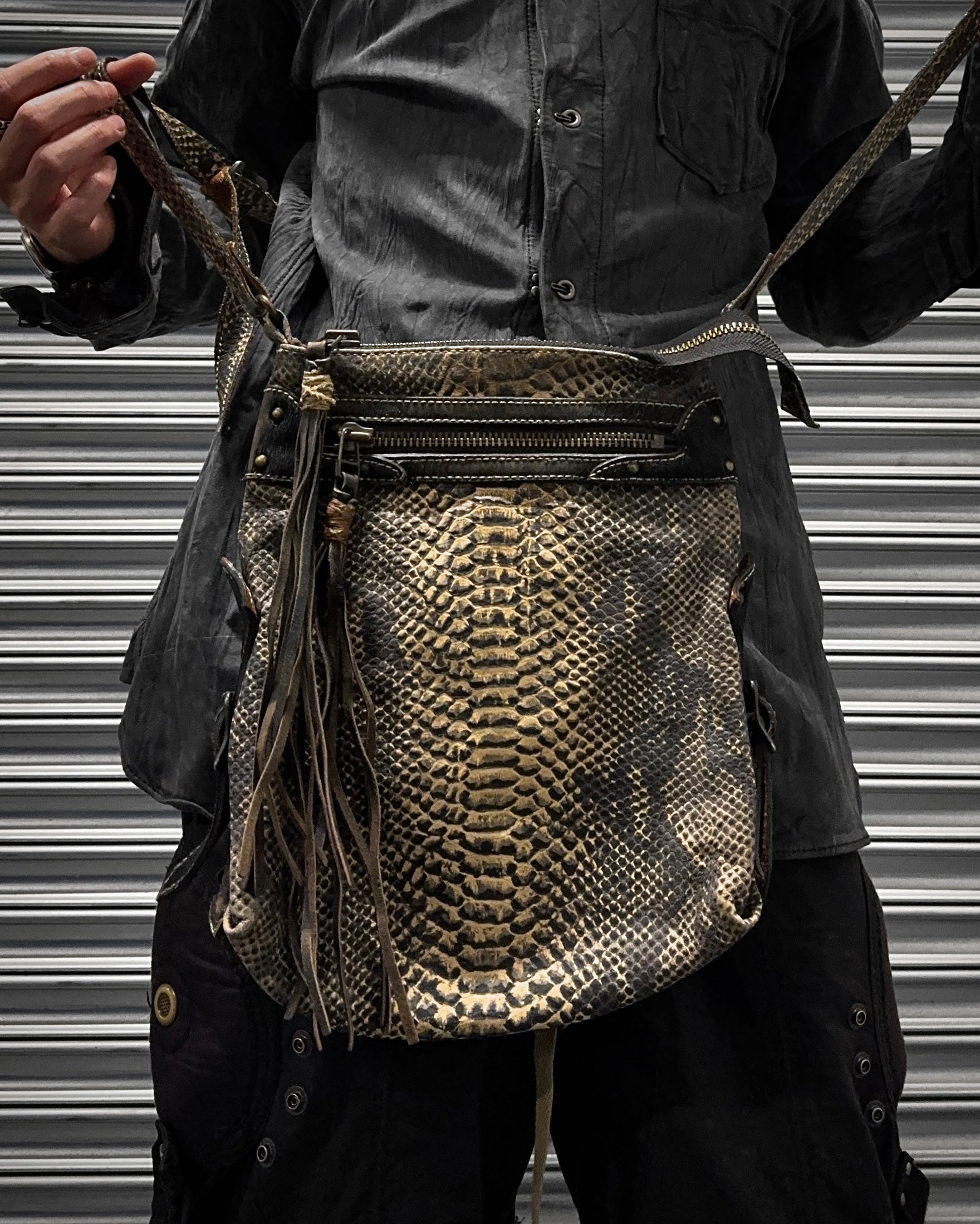 A person in dark clothing holds a Kyoji Maruyama Early 00s Yellow Crocodile Embossed Leather Side Bag, standing in front of a metal shutter.