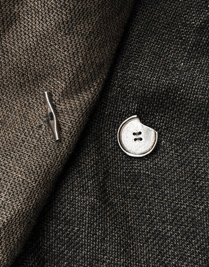 A silver “coin” button with a missing section and a matching toggle accent the textured fabric of the TaichiMurakami “Coin” Taped Seam Collarless Work Blazer, embodying the brand’s industrial minimalism.
