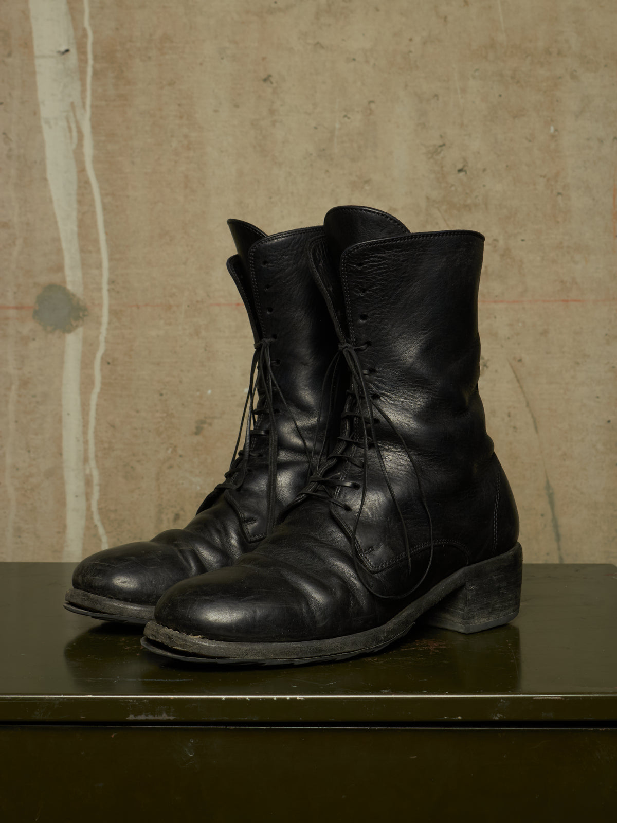 A pair of black leather lace-up combat boots with a rugged, scuffed finish, displayed on a dark green surface against a textured beige wall.