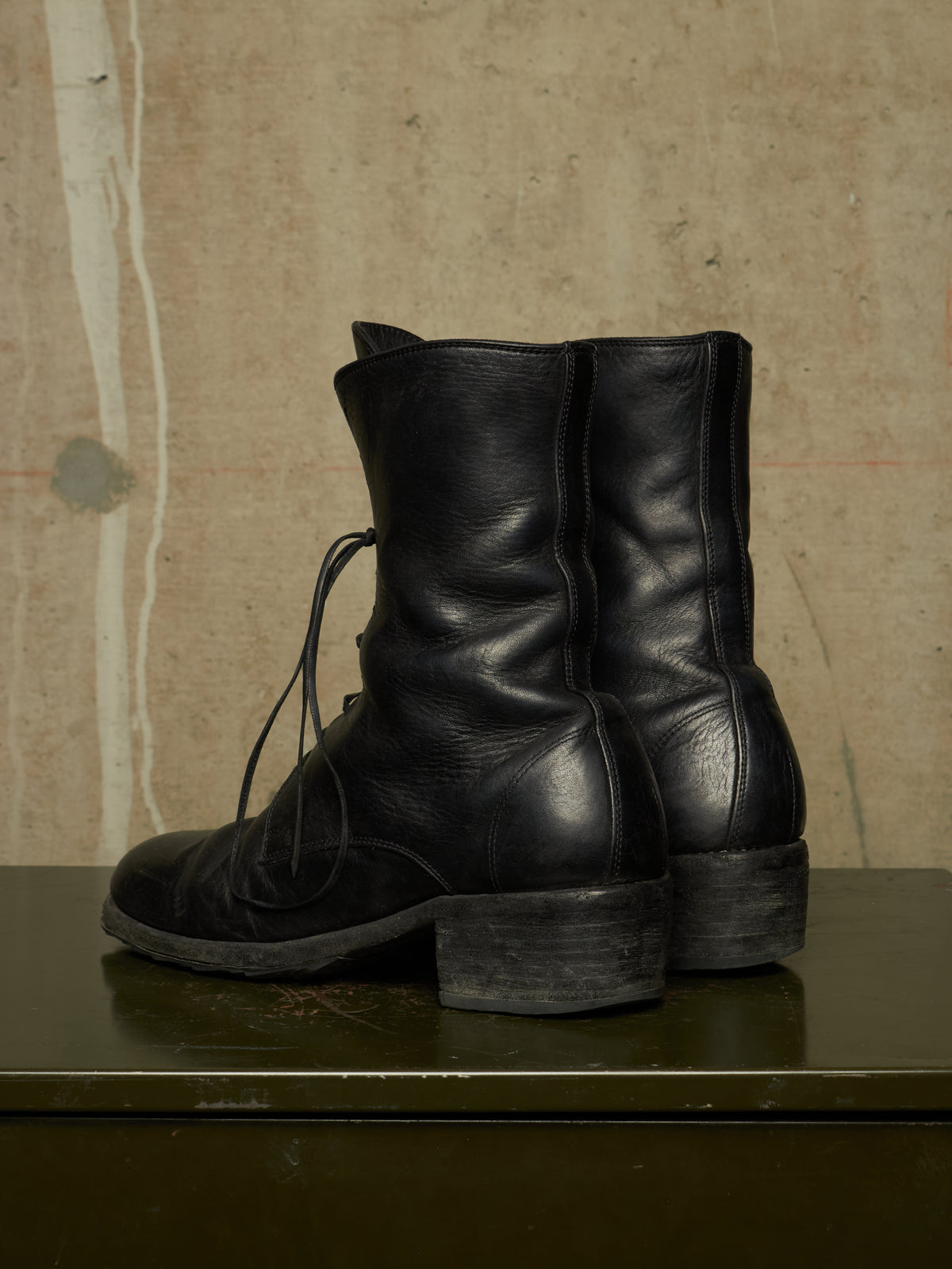 A pair of black leather lace-up ankle boots with chunky block heels, displayed on a dark green surface against a textured beige concrete wall.