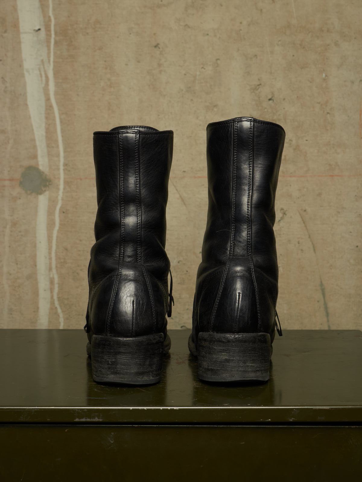 A pair of black leather mid-calf boots shown from the back, resting on a dark green surface against a textured beige wall.