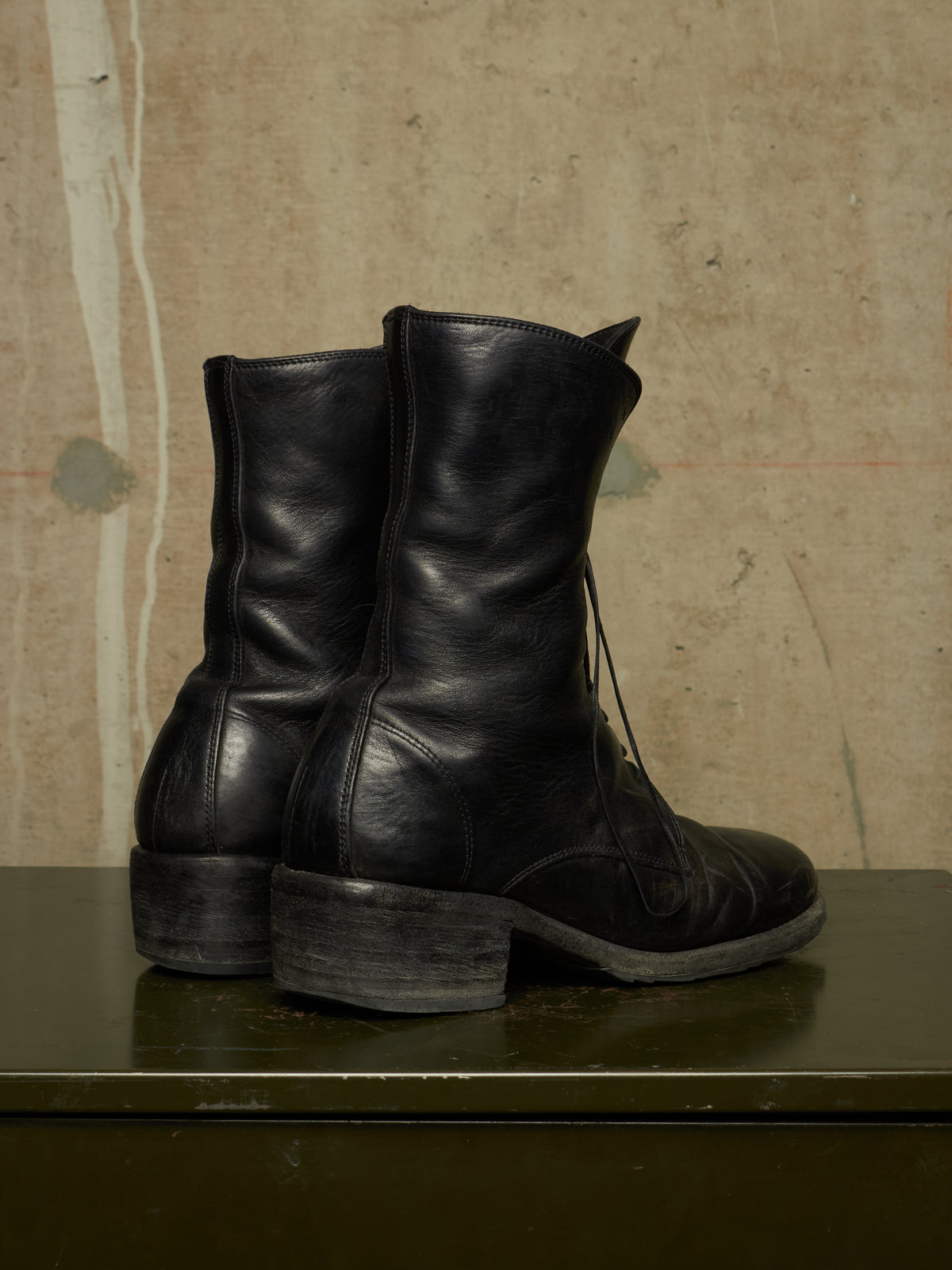 A pair of black leather lace-up ankle boots with low block heels, shown from the side and back on a dark green surface against a textured beige wall.