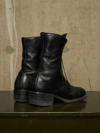 A pair of black leather lace-up ankle boots with low block heels, shown from the side and back on a dark green surface against a textured beige wall.