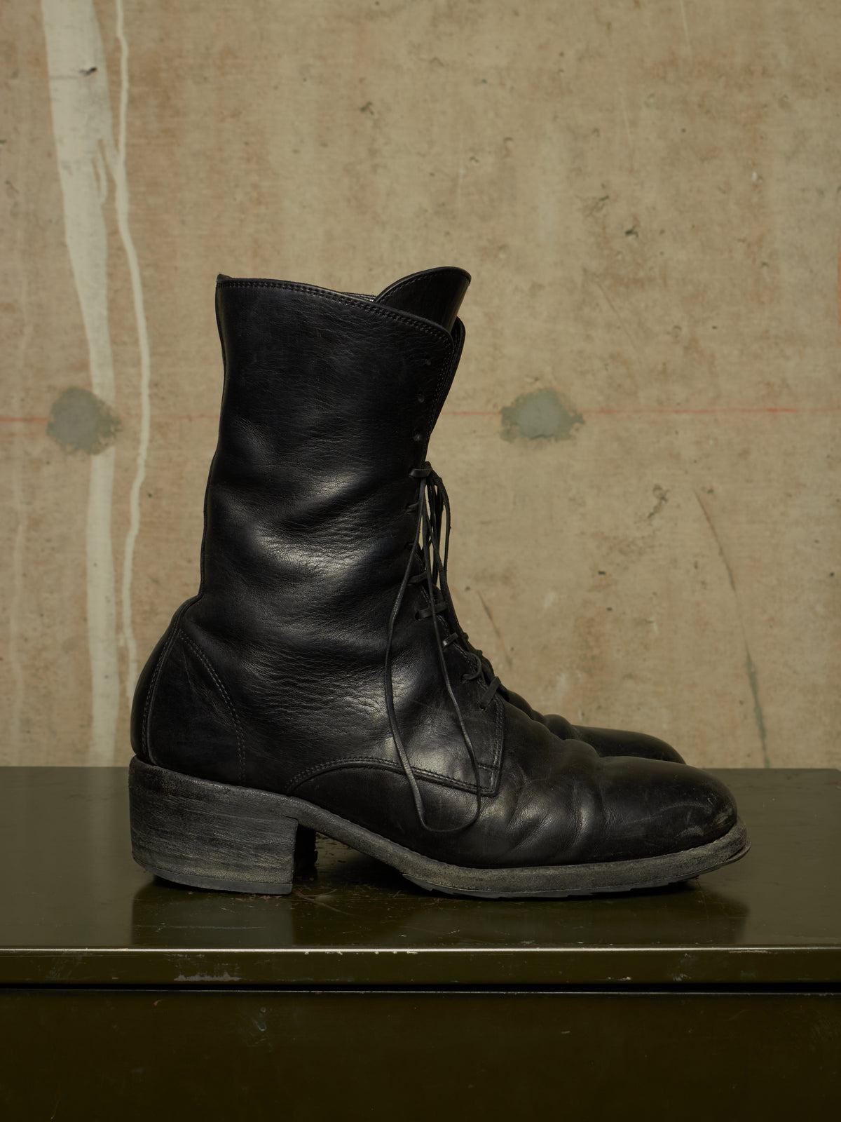 A pair of black leather lace-up ankle boots with a low block heel, displayed on a dark green surface against a textured beige concrete wall.