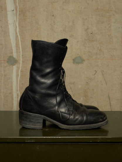 A pair of black leather lace-up ankle boots with a low block heel, displayed on a dark green surface against a textured beige concrete wall.