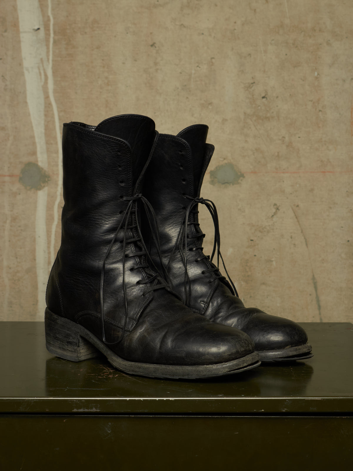A pair of black leather lace-up combat boots with a rugged low heel, displayed on a dark green metal surface against a textured beige background.