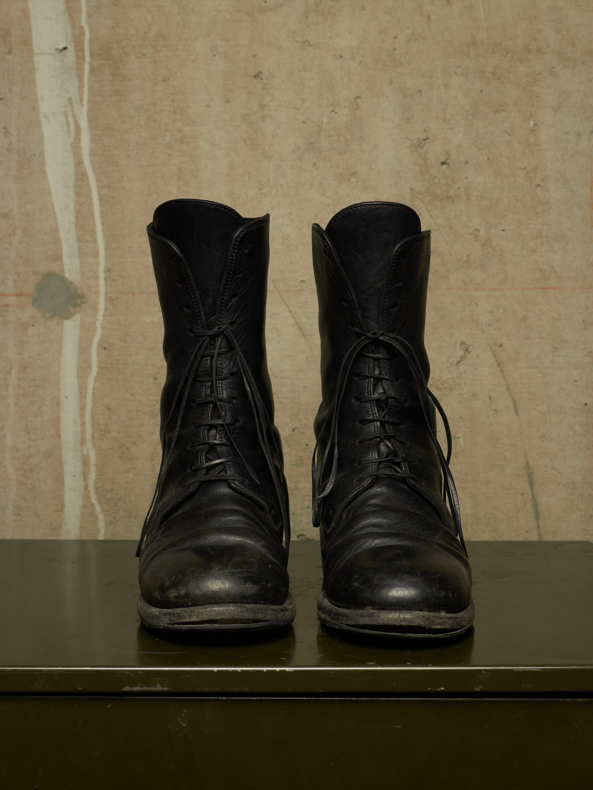 A pair of worn black leather lace-up ankle boots displayed side-by-side on a dark green surface against a textured beige wall.