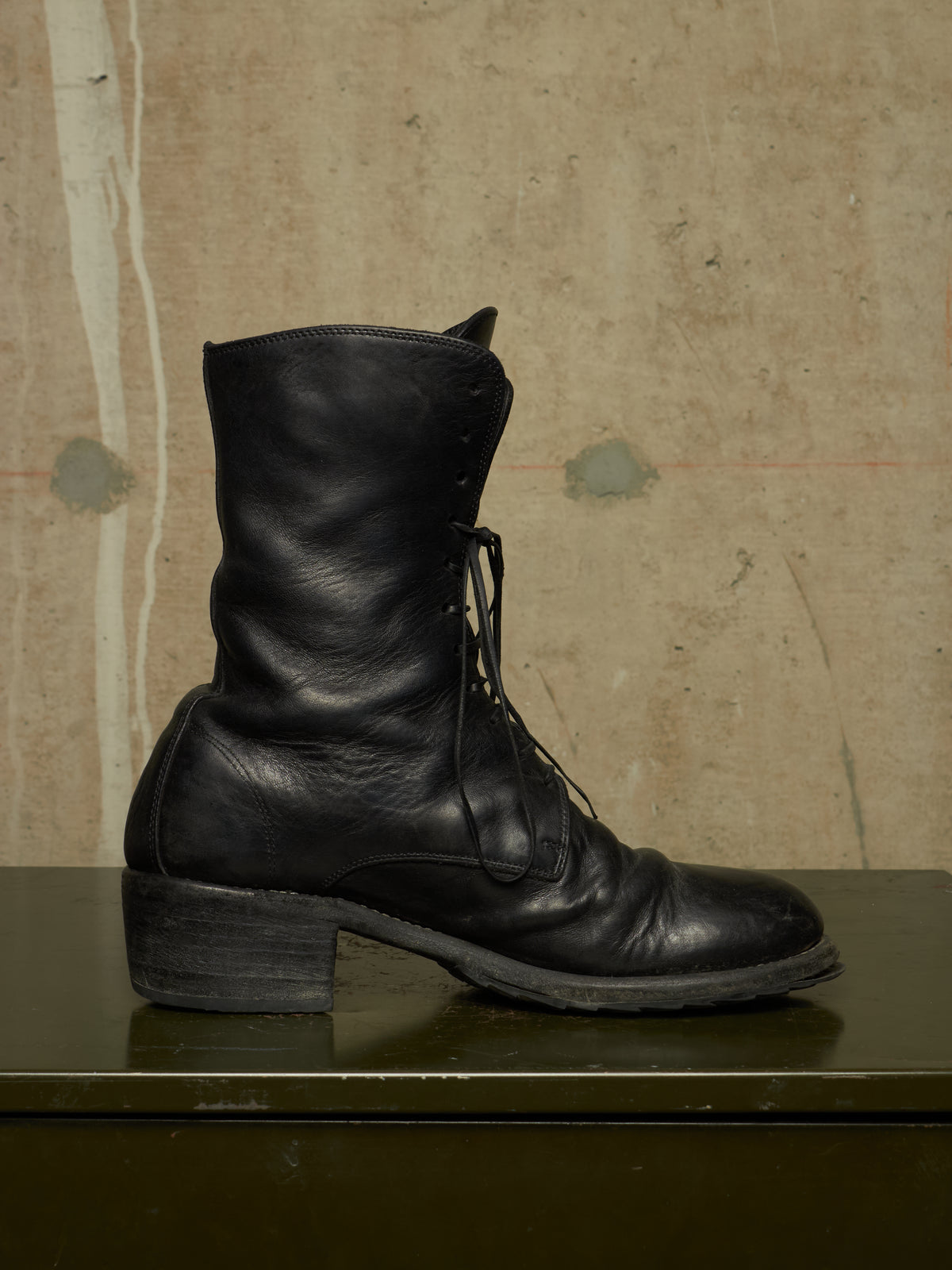 A single black leather lace-up boot with a low block heel, resting on a dark green surface against a textured beige concrete wall.