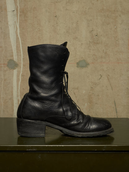 A single black leather lace-up boot with a low block heel, resting on a dark green surface against a textured beige concrete wall.