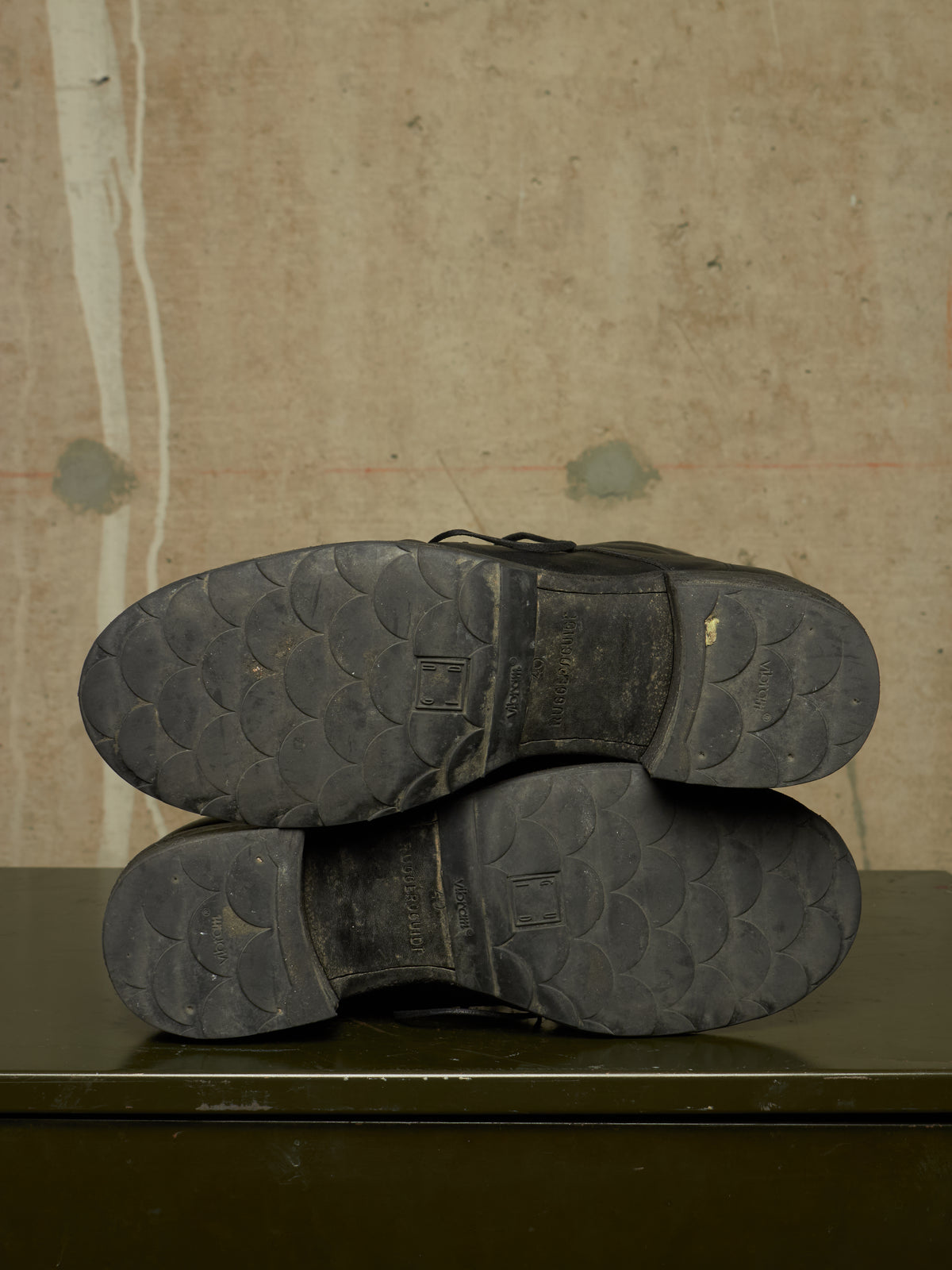 A pair of black leather boots shown from the sole side, resting on a dark green surface against a textured beige wall.