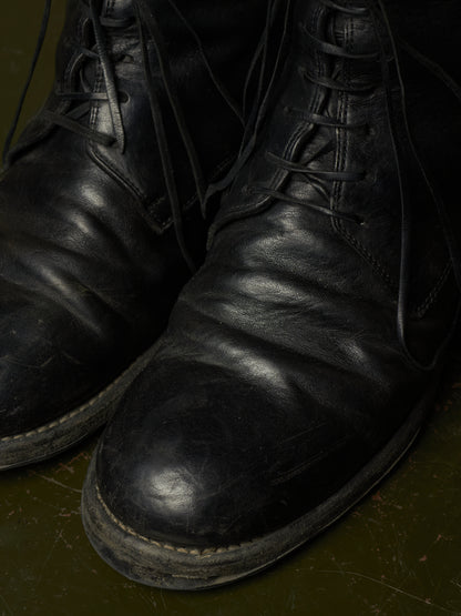 A close-up shot of a pair of worn black leather lace-up boots resting on a dark olive green floor.
