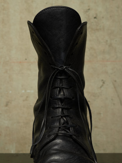 A close-up of a black leather lace-up boot against a textured beige background.
