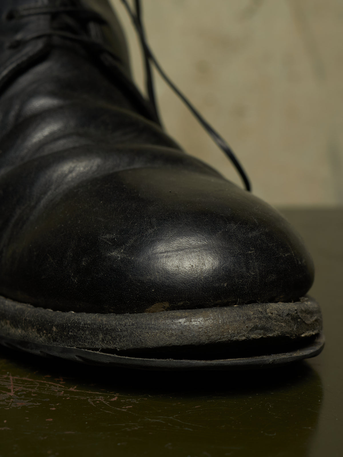 A close-up shot of a worn black leather lace-up boot, showing significant scuffing and damage along the toe and sole, resting on a dark reflective surface.