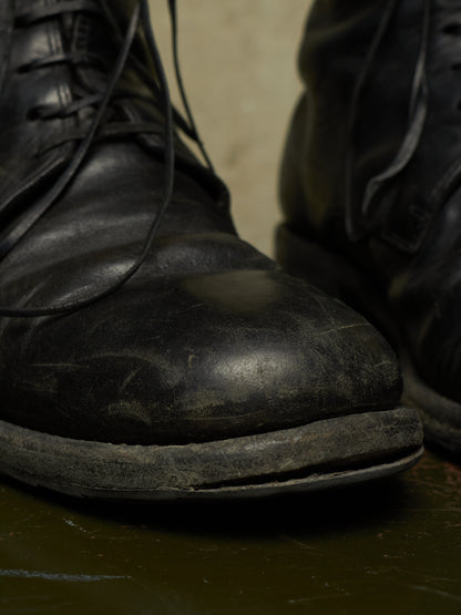 A close-up shot of a worn black leather lace-up boot with a visibly cracked sole, sitting on a dark reflective surface with another boot partially visible in the background.