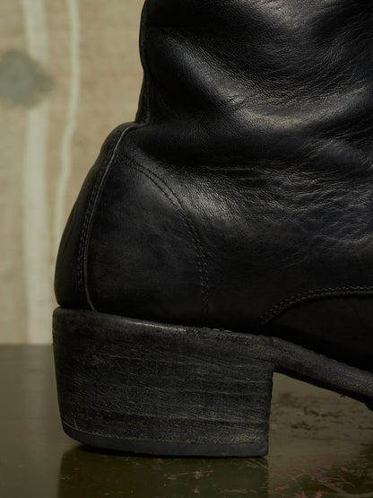 Close-up of a black leather boot showing the heel, side seam, and textured leather surface, photographed against a softly blurred neutral background.